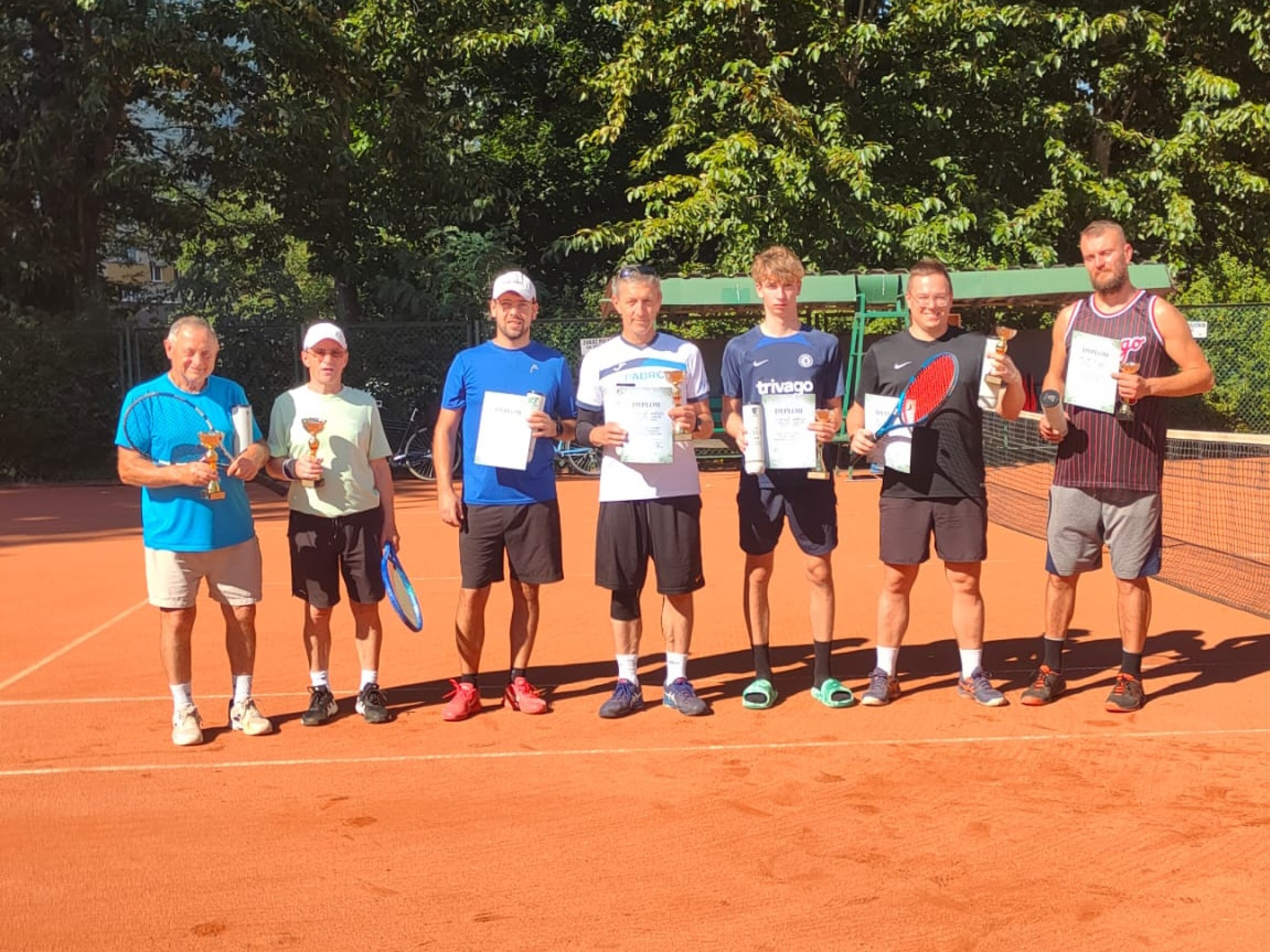 Laureaci turnieju stojący na korcie i trzymajacy w reku dyplomy, pilki i puchary.